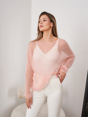 Woman wearing a light pink knitted sweater and white pants, standing with one hand on her hip. She...