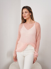 Woman in a soft pink knitted sweater and white pants, stands in a relaxed pose. Her long brown hair...