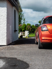 Fototapeta premium Small sport red car parked by a house with white walls, rural area in the background. Warm sunny day. Living in a country side concept. Commute and transportation theme.
