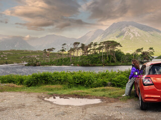 Girl in blue jacket standing by red car and looking at lake with Twelve pines island and amazing...
