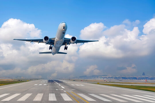 View airplane ascends into the sky after leaving the runway. The clear blue sky is dotted with white clouds, and an airport is visible in the background, showcasing a bustling travel scene