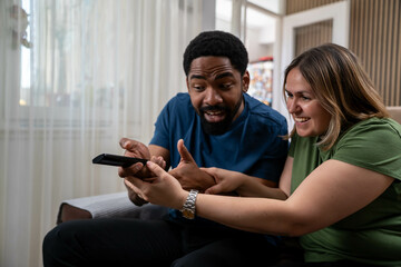 Interracial couple playfully fighting over TV remote control at home, smiling and enjoying time...