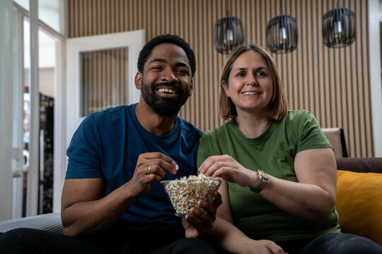 Interracial couple smiling and eating popcorn while watching TV together at home, enjoying happy moment in cozy living room, joyful everyday lifestyle and connection - Powered by Adobe