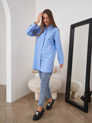 Young woman with long hair in a blue shirt and jeans, touching her head, stands indoors near a...