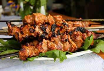 Close-up of juicy grilled pork skewers served on a white plate with fresh oak leaves. Rustic table setup, ideal for a traditional outdoor barbecue feast.