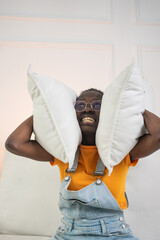 Frustrated young african american woman covering her ears with pillows while sitting on bed,...