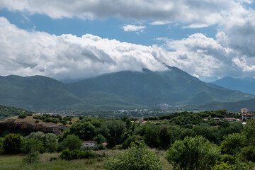 A serene panoramic view of lush green hills and a rural village nestled in a valley with dramatic clouds rolling over a mountain range in the background.