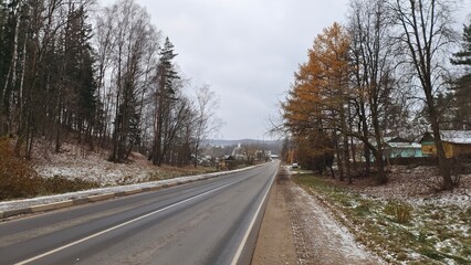 The asphalt road with markings passes among yellowed larches and other trees. On the sandy roadsides lies the first snow. The road leads to the houses of the settlement. Overcast autumn weather