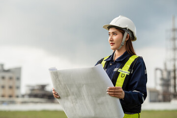 Female Engineer in Uniform Reviewing Blueprints at an Industrial Plant. A woman wearing a yellow...