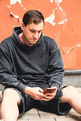 On a sunny day a young man wearing a gray hoodie and denim shorts is sitting on the pavement...