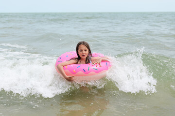 A surprised young girl with long hair, in a pink donut-shaped inflatable ring, reacts to a wave...