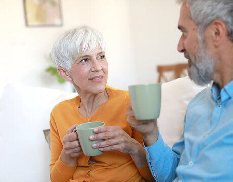 Portrait of a happy senior couple embracing talking and drinking coffee or tea at home - Powered by Adobe