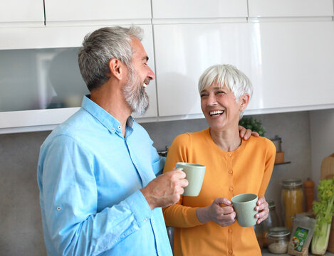 A man and a woman are smiling and holding cups of coffee in a kitchen. Scene is happy and friendly