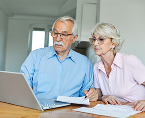 Senior couple checking bills and calculating expenses using a laptop at home, debt taxes concept,...
