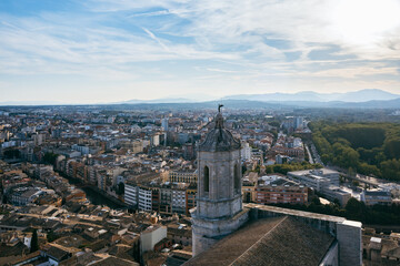 Obraz premium An aerial view of Girona's historic city center, featuring the cathedral, the Onyar River winding through colorful buildings, and ancient city walls amidst lush hills
