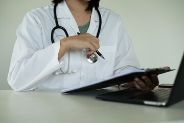Young asian woman doctor in white uniform with stethoscope taking notes, using laptop, mobile phone, writing in medical journal, professional therapist practitioner filling documents or patient card.