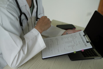 Young asian woman doctor in white uniform with stethoscope taking notes, using laptop, mobile phone, writing in medical journal, professional therapist practitioner filling documents or patient card.