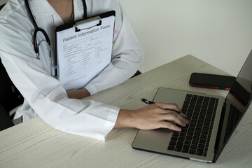 Young asian woman doctor in white uniform with stethoscope taking notes, using laptop, mobile phone, writing in medical journal, professional therapist practitioner filling documents or patient card.