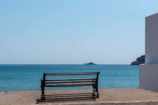 Old empty wooden bench facing calm blue sea with a distant yacht and rocky island under a vast clear sky - Powered by Adobe