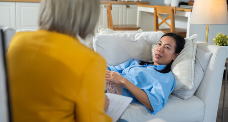 Obraz premium Senior Asian female psychologist attentively taking notes while listening to an Asian woman patient lying on a sofa during a therapy session, providing mental health support and counseling.