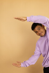 Vertical image of a young brown skinned Latino man wearing a lilac sweatshirt pointing with both hands at a copy space on a light brown background, advertising space to promote