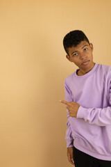 Vertical image of a young brown skinned Latino man wearing a lilac sweatshirt pointing with his fingers at a copy space on a light brown background, advertising space to promote
