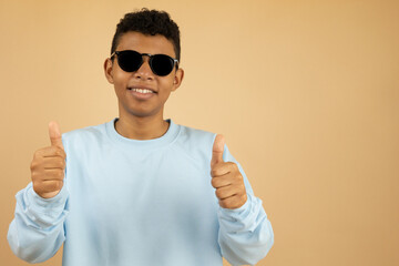 Smiling young Latino man wearing dark glasses and a pastel blue sweatshirt making a thumbs up gesture with both hands on a light brown background with copy space