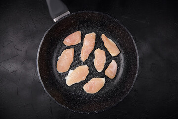 Raw chicken breast pieces arranged in a frying pan on a dark textured surface, showcasing the...