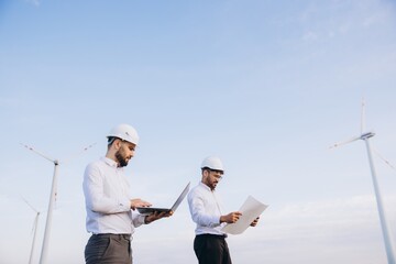 Engineers working on a laptop and checking blueprints next to wind turbines