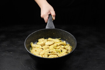 Hand holding frying pan with cooked chicken pieces, seasoned and sauteed, on a dark background,...