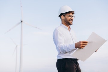 Smiling engineer holding blueprint inspecting wind turbines in field