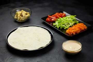 Fresh ingredients arranged on a dark surface, featuring a tortilla, sliced vegetables, and a bowl...