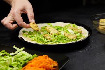 Hand placing crispy chicken pieces onto a fresh bed of lettuce in a black plate, surrounded by vibrant shredded carrots and leafy greens, showcasing a healthy meal preparation