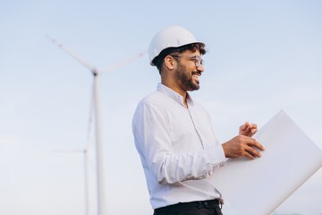 Smiling engineer holding blueprint inspecting wind turbine in field