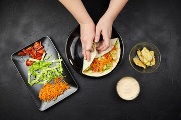 Hands of a person skillfully preparing a fresh vegetable wrap with colorful ingredients on a black textured surface, showcasing culinary creativity and healthy eating
