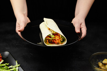 Freshly prepared wrap filled with colorful vegetables and greens is being served on a black plate,...