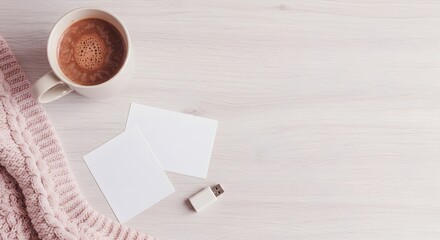 A mug of hot coffee, a pink knit scarf, blank cards, and a usb drive are arranged on a wooden table for a cozy scene