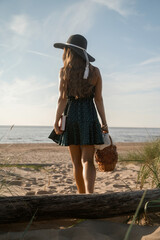 A woman in a black hat and green polka dot dress walks barefoot on sandy dunes, carrying a red book and a wicker basket, with the sea in the background