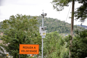 Radar fixo com placa “Reduza a Velocidade” em estrada cercada por vegetação. Imagem sobre segurança no trânsito e controle de velocidade em áreas rurais.