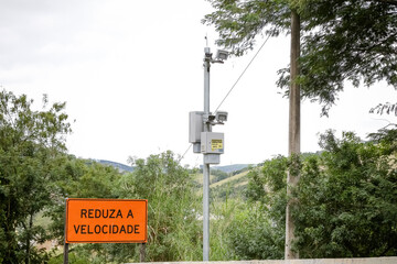 Radar fixo com placa “Reduza a Velocidade” em estrada cercada por vegetação. Imagem sobre...