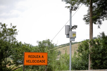 Radar fixo com placa “Reduza a Velocidade” em estrada cercada por vegetação. Imagem sobre...