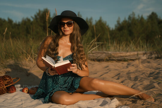 A woman in a wide-brimmed hat and green polka-dot dress holds a feather and a book, smiling serenely on a sunlit beach