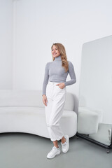 Smiling woman with long hair in a gray turtleneck and white pants stands next to a full-length...