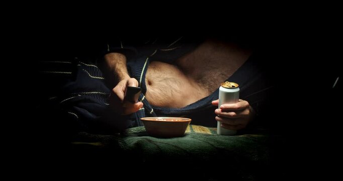 A sloppy adult man in a dressing gown with a bare belly and a TV remote control lying on a sofa eats chips and drinks a drink from a can. A forty-year-old skuf