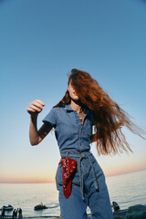 Lifestyle woman in modern boho-western denim street style stands by the sea with wind-blown red...