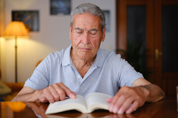 Front view of an old man reading a book and pointing at a page in his living room.