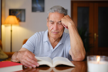 Elderly man reading a book at home with a lit candle on the table, resting his head on one hand with a thoughtful expression.
