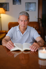 Vertical image of an old man turning the page of a book next to a candle with his reflection on the table.