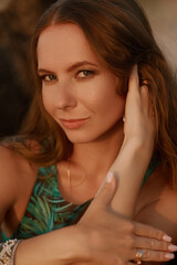 A woman in a green palm-print bikini, adorned with jewelry, gazes softly into the distance,...