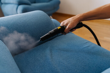 Cleaning service worker holding steam cleaner machine and cleaning blue sofa, removing dirt and...